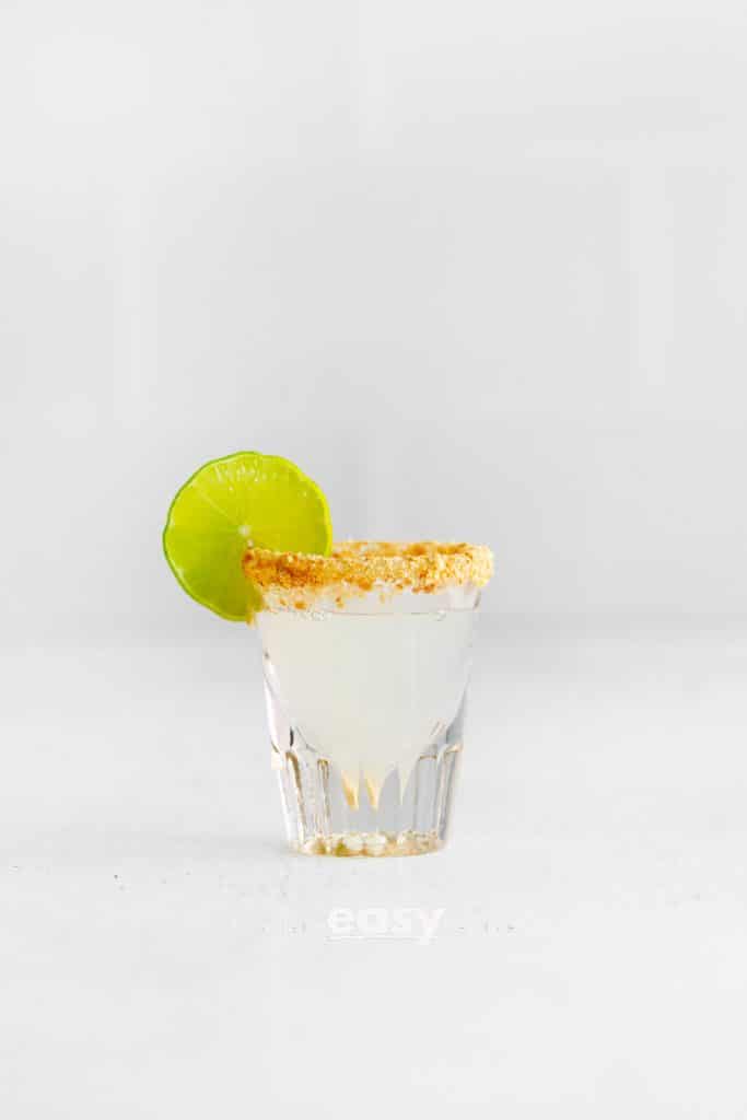 clear shot glass with a milky white clear liquid. Brown crumbled graphams on rim of shot glass. Lime wheel on top side of glass. L