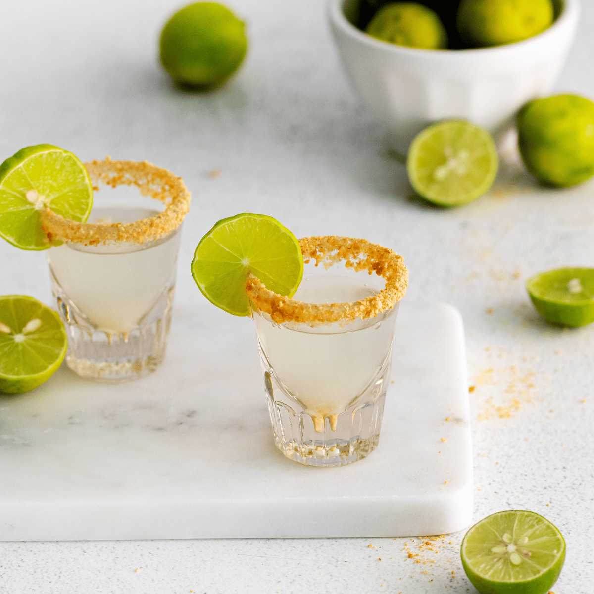 clear shot glass with a milky white clear liquid. Brown crumbled graphams on rim of shot glass. Lime wheel on top side of glass. Limes in backaground