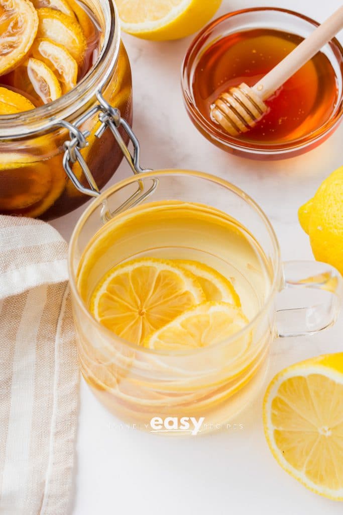 clear tea cup with clear water and lemon slices. Also visible is a honey jar with scooper and halved lemons