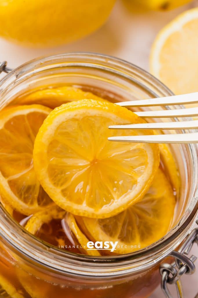 clear jar with lid. Inside are sliced lemons in brown dark honey. slices halved lemons on side and above and honey jar also visable with fork getting a lemon slice from jar