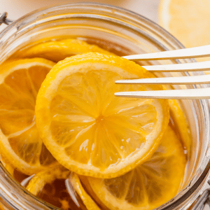 clear jar with lid. Inside are sliced lemons in brown dark honey. slices halved lemons on side and above and honey jar also visable