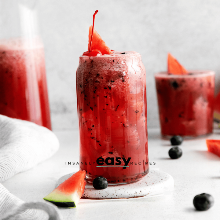 clear glass filled with crushed ice and red liquid with black flecks. Watermelon, cherry and blueberries on white table with 3 glasses filled with that liquid