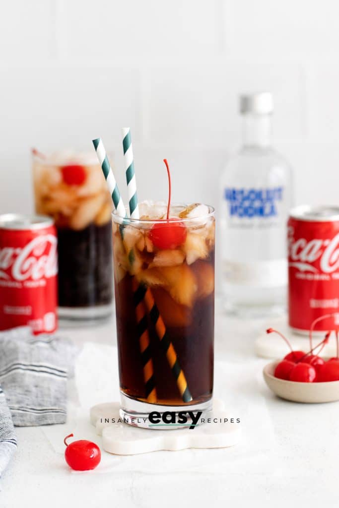 two tall glasses with brown liquid, ice cubes, cherry and straws. In background are two coke can and a vodka bottle. A cherry and bowl of cherries on table