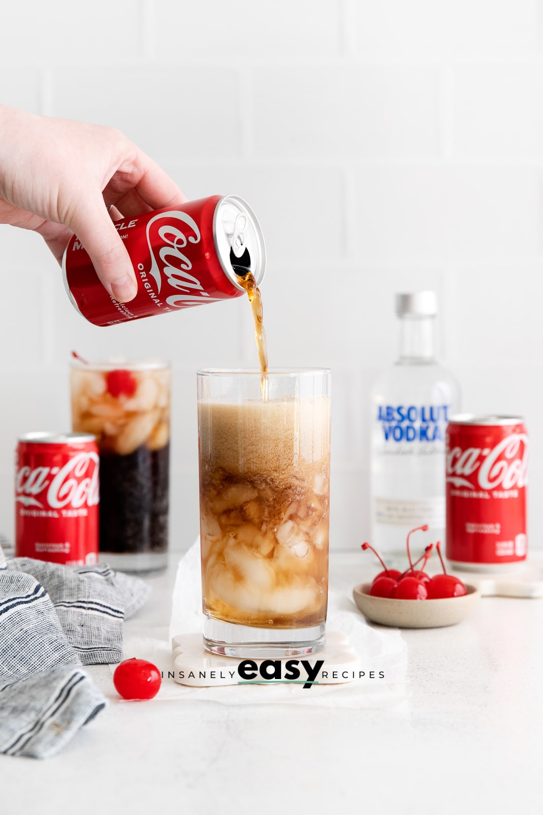two tall glasses with brown liquid, ice cubes, cherry and straws. In background are two coke can and a vodka bottle. A cherry and bowl of cherries on table
