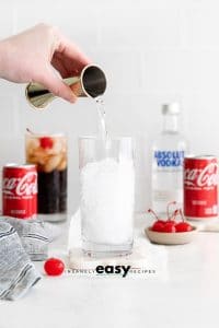 pouring a clear liquid into a glass with coke and vodka bottles behind it