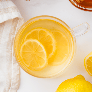 clear tea cup with clear water and lemon slices. Also visible is a honey jar with scooper and halved lemons