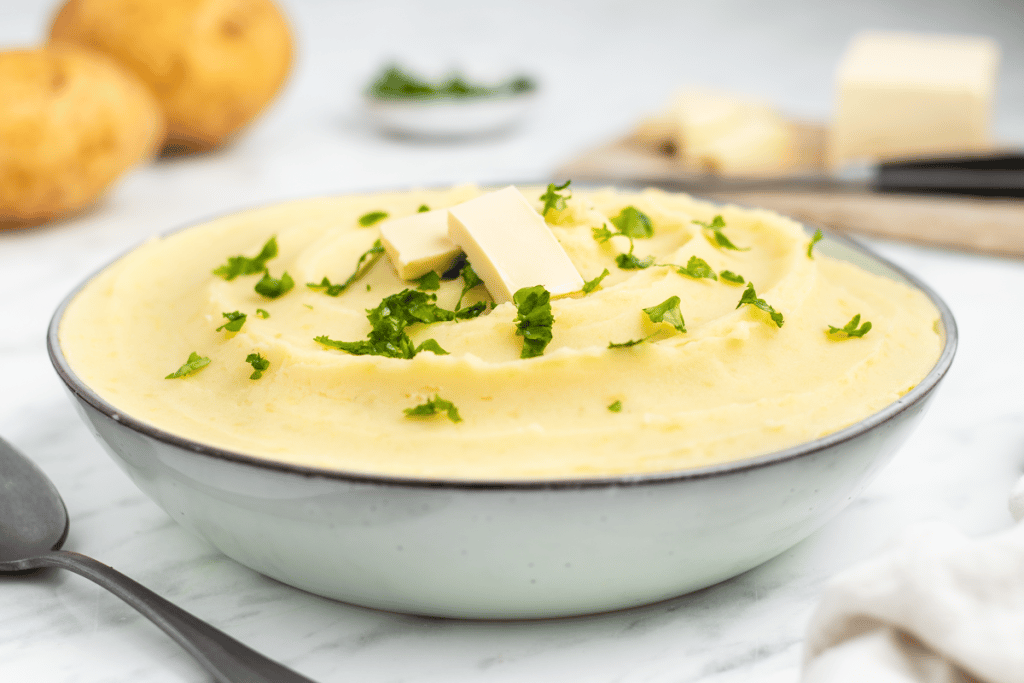 white bowl with mashed potatoes, butter slabs and fresh parsley on top. White marble like counter, potatoes in top left butter in top right