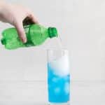 hand pouring sprite into a tall glass that already has blue liquid