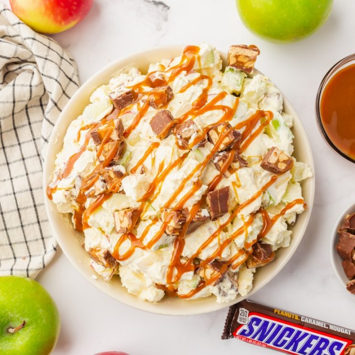 Snicker Apple Salad in a white bowl. Looks like fluffy white stuff, with apple chunks, snickers chunks, marshmallows and caramel sauce,