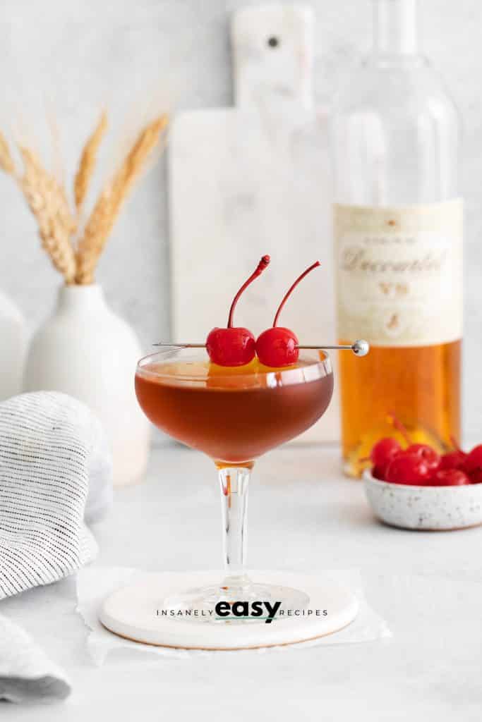 pictured is a brandy manhattan. It is a brown liquid drink with two cherries on it. With a bottle of brandy in the top right and cherries in a bowl on the righ