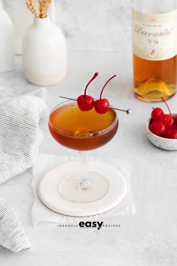 pictured is a brandy manhattan. It is a brown liquid drink with two cherries on it. With a bottle of brandy in the top right and cherries in a bowl on the right
