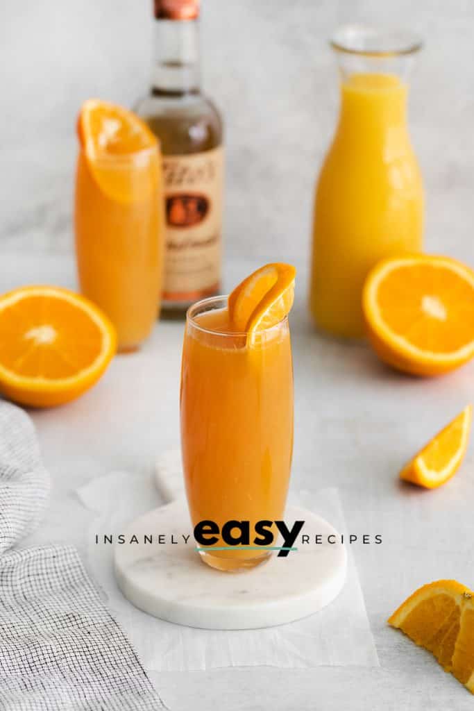 two tall glasess of manmosa. Orange liquid in two tall glasses with orange wheel on top. Sliced orange to the side, along with a vodka bottle and an jar of orange juice