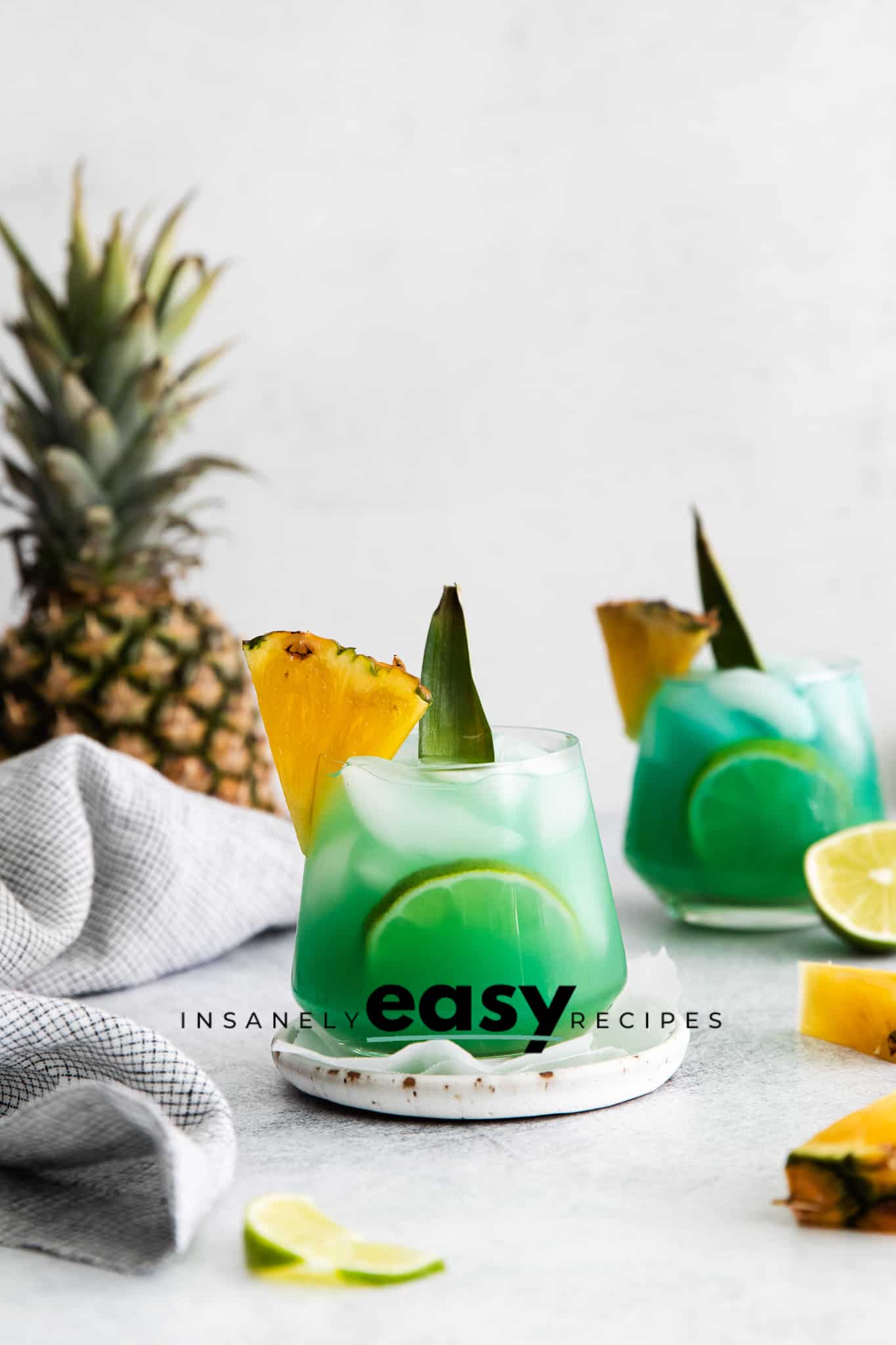 Pictured is a mermaid water. There are two short glasses, filled with green/blue liquid and ice cubes. There is a pineapple wedge and leaf on each rim. There are pineapple pieces and lime pieces on the table with a pineapple in the back left.