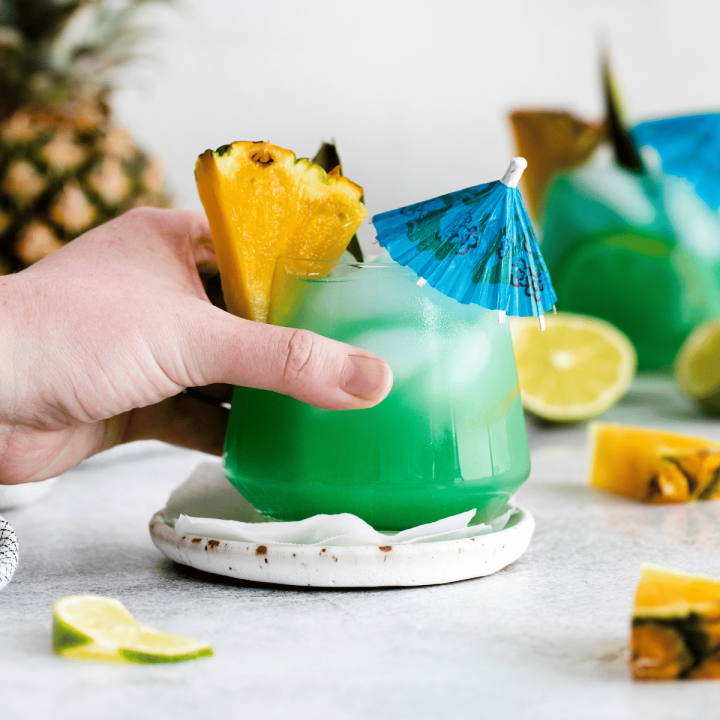 Pictured is a mermaid water. There are two short glasses, filled with green/blue liquid and ice cubes. There is a pineapple wedge and leaf on each rim. There are pineapple pieces and lime pieces on the table with a pineapple in the back left. a hand holding the glass in front