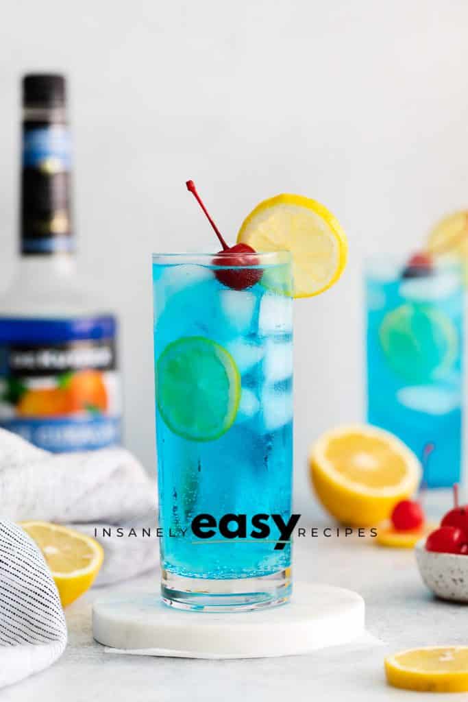 tall clear glass with blue liquid and ice cubes A lemon wheel on top and a cherry two glasses ike that and a bottle of blue alcohol in the back