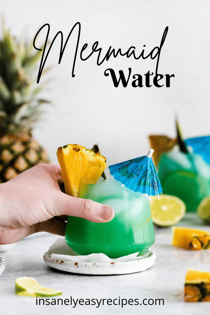 Pictured is a mermaid water. There are two short glasses, filled with green/blue liquid and ice cubes. There is a pineapple wedge and leaf on each rim. There are pineapple pieces and lime pieces on the table with a pineapple in the back left. with text overylay: mermaid water
