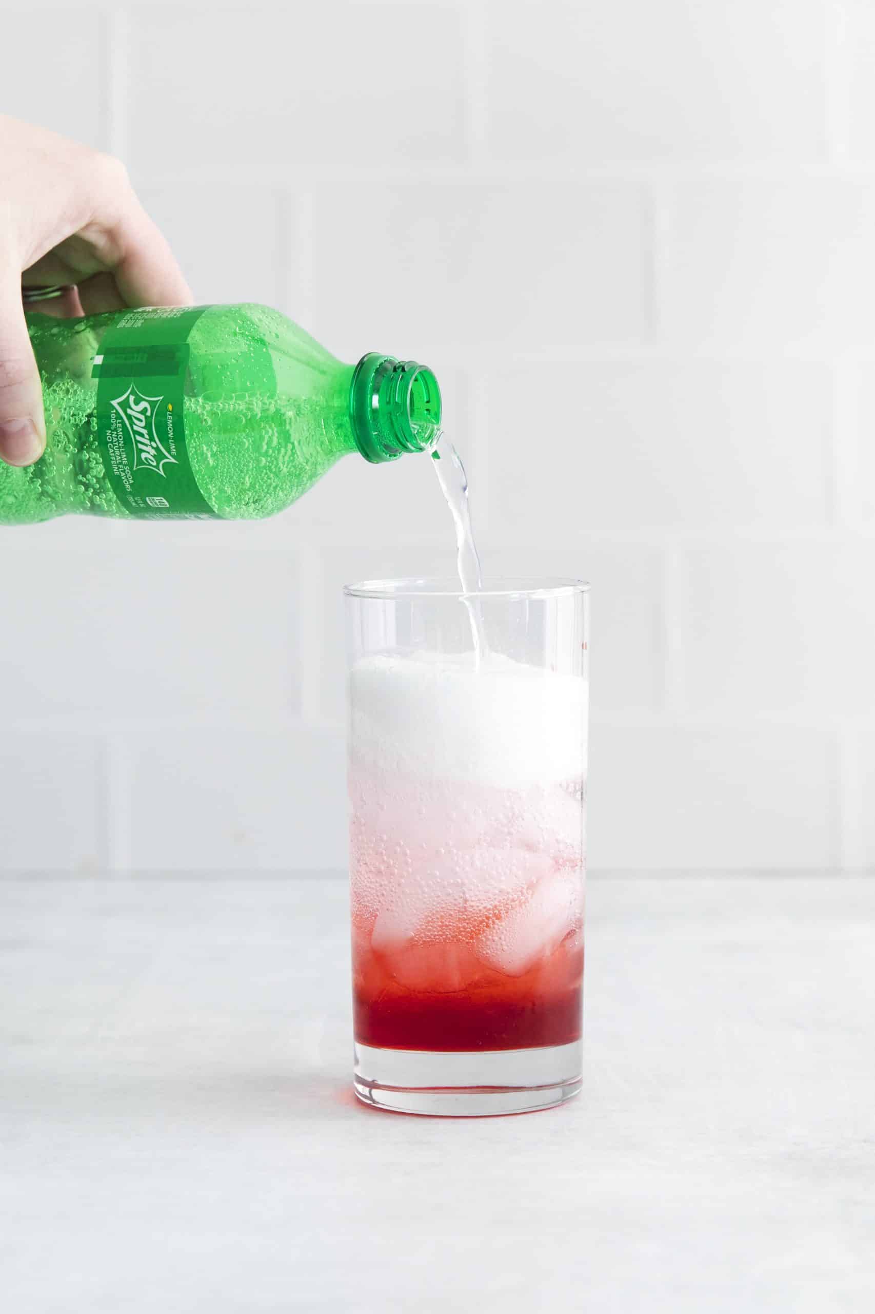 Tall glass with red liquid and sprite being poured into it