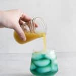 yellow liquid being poured into a glass with green liquid and ice cubes