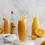 two tall glasess of manmosa. Orange liquid in two tall glasses with orange wheel on top. Sliced orange to the side, along with a vodka bottle and an jar of orange juice
