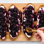 top view photo of 4 toasted sourdough bread slices with yogurt and blueberry jam spread on top, and a hand drizzling honey on top of each slice, on a wooden cutting board