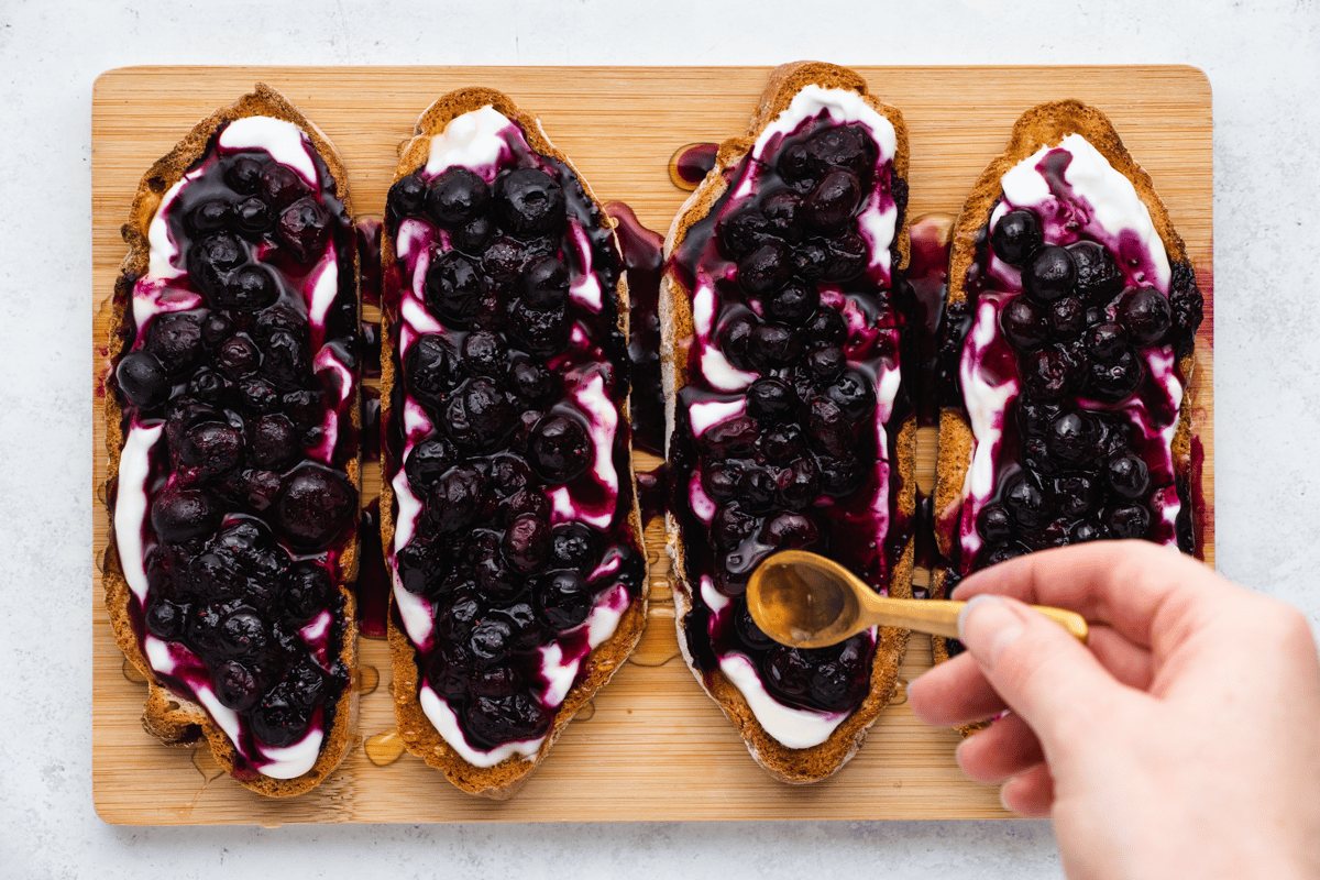 top view photo of 4 toasted sourdough bread slices with yogurt and blueberry jam spread on top, and a hand drizzling honey on top of each slice, on a wooden cutting board