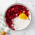 top view photo of cranberry jam ingredients (cranberries, sugar, water, and lemon zest) in a saucepan with a towel next to it