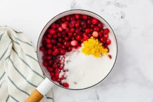 top view photo of cranberry jam ingredients (cranberries, sugar, water, and lemon zest) in a saucepan with a towel next to it