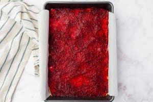 top view photo of cranberry lemon bars with a layer of crust and cranberry jam spread on top, in a pan, with a towel next to it