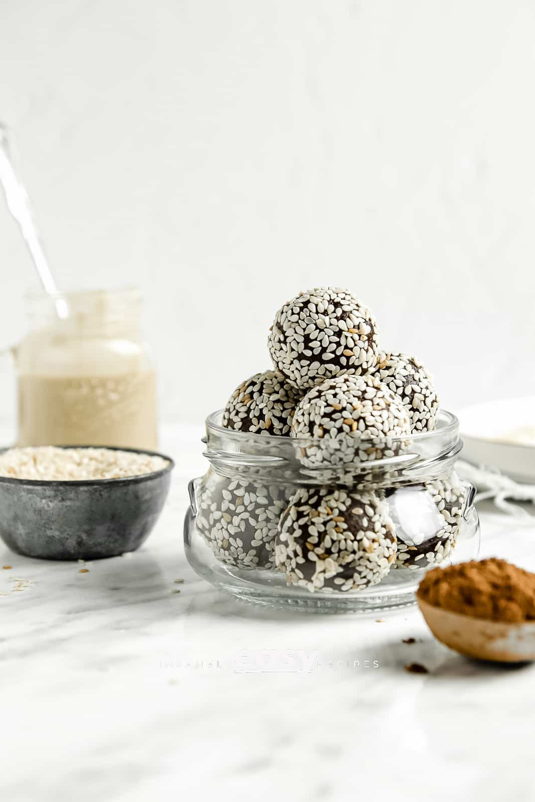 photo of bliss balls in a clear canning jar, with a spoon filled with cocoa powder in the foreground and a small silver bowl of almond flour, a mason jar of tahini paste with a spoon in the jar, and a white plate in the background