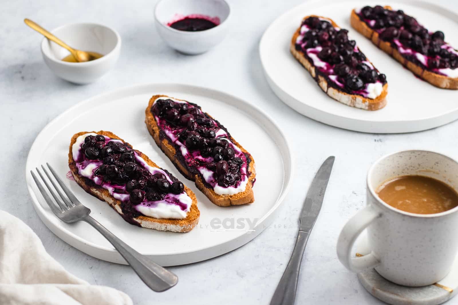 photo of 2 white plates with 2 servings of blueberry toast on each plate. there is also a fork, knife, and mug of coffee in the foreground, and a small white bowl of honey and blueberry jam in the background