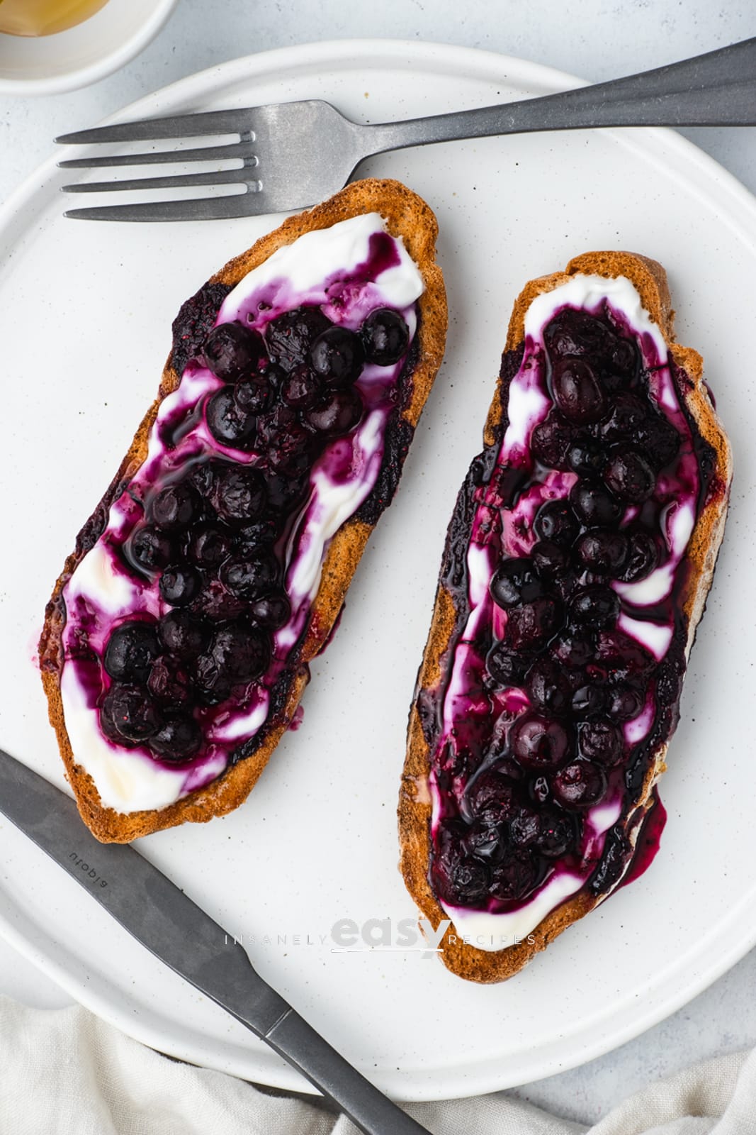 top view photo of 2 slices of blueberry toast on a white plate with a fork and small bowl of honey above it, and a butter knife and white kitchen towel below it
