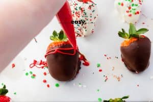 person decorate brown chocolate covered strawberry with red pipping