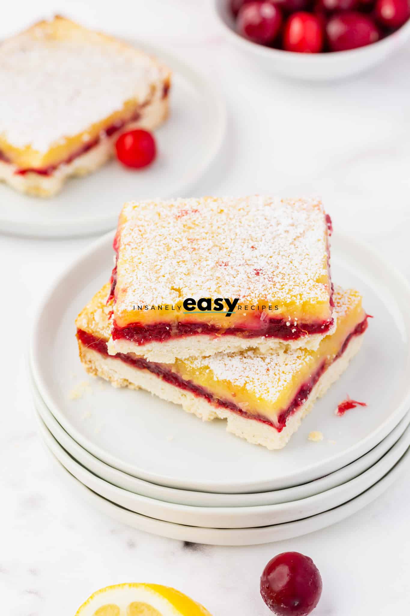 photo of two cranberry lemon bar slices stacked on top of eachother on a stack of white plates with a slice of cranberry lemon bar in the background on a white black with a decorative cranberry next to it