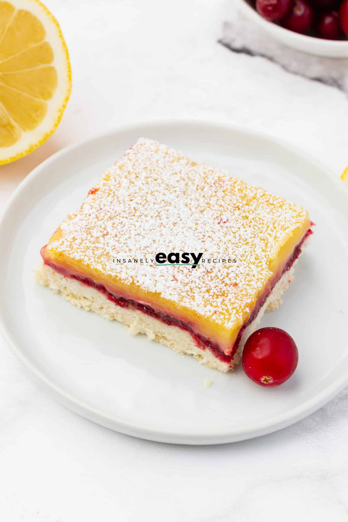 photo of a slice of cranberry lemon bars on white plate with a decorative cranberry next to it and a lemon slice in the background
