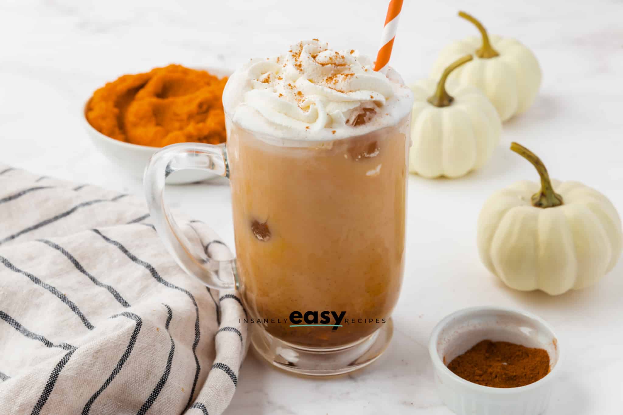 pumpkin spice latte in a clear mug with pumpkin puree in the back left and white pumpkins in the back top right.