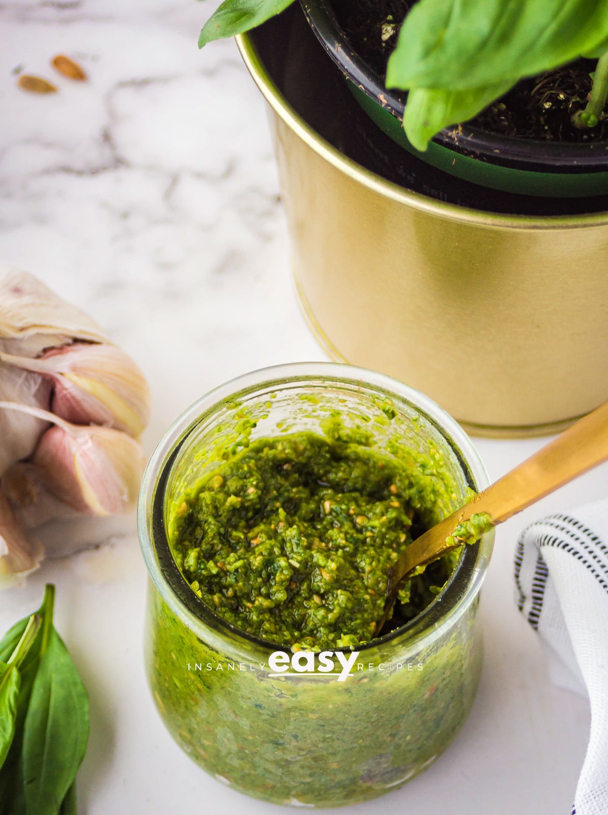 photo of nut free pesto in a small glass jar with a gold spoon dipped inside. the jar is surrounded by basil, garlic cloves, a white kitchen towel, and a gold bowl