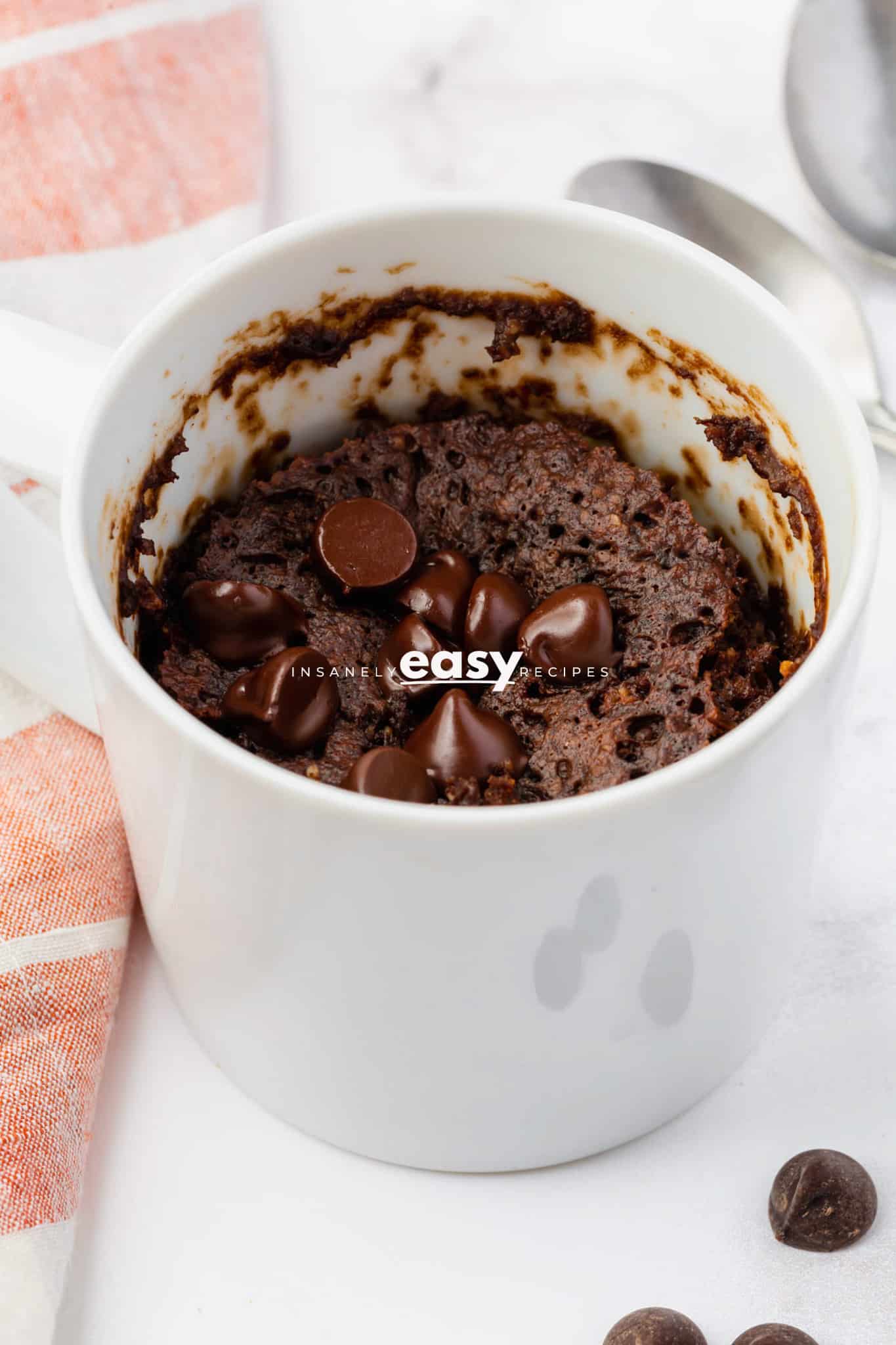 photo of chocolate protein mug cake in a white mug surrounded by a couple chocolate chips, a towel, and 2 spoons
