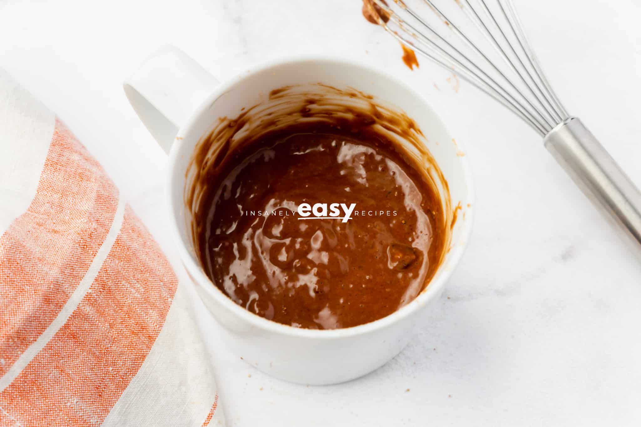 top view photo of a mug with batter to make protein mug cake with a kitchen towel and a wire whisk next to it