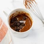 top view photo of a mug with batter and chocolate chips to make protein mug cake with a kitchen towel and a wire whisk next to it