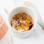 top view photo of a mug with wet and dry ingredients to make protein mug cake with a kitchen towel and a wire whisk next to it