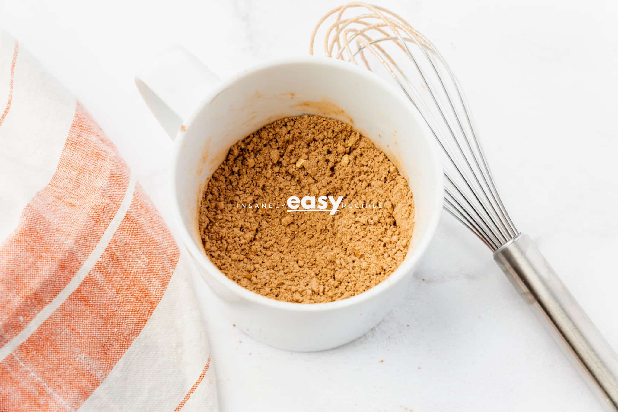 top view photo of a mug with dry ingredients to make protein mug cake with a kitchen towel and a wire whisk next to it