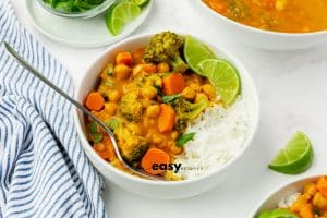 White bowl, filled with rice and pumpkin curry. You can see chickpeas and carrots in a sauce. There are limes cut into quarters and a blue/white striped napkin to the left.