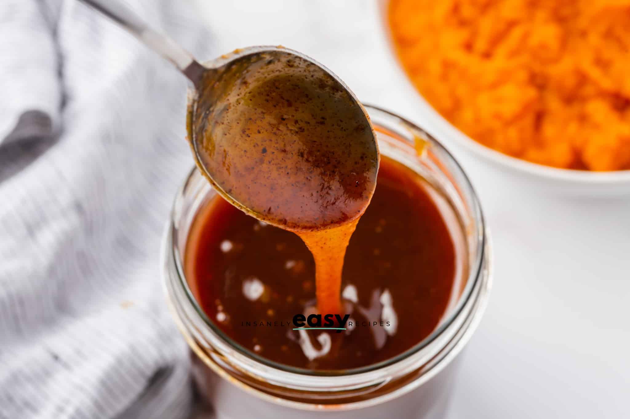 glass jar with brown liquid, metal spoon dipping into the liquid and some pouring off spoon. Top right corner has puree pumpkin in a white bowl. Gray napkin to left side.