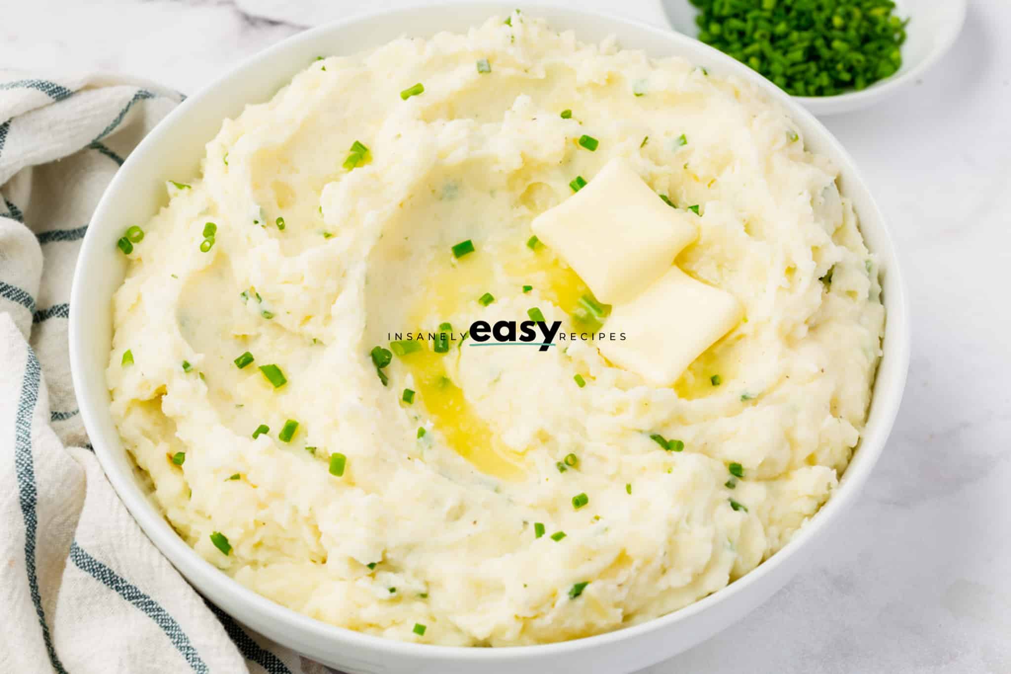 creamy ranch mashed potatoes in a white bowl. Potatoes have green seasoning on top and two butter pads