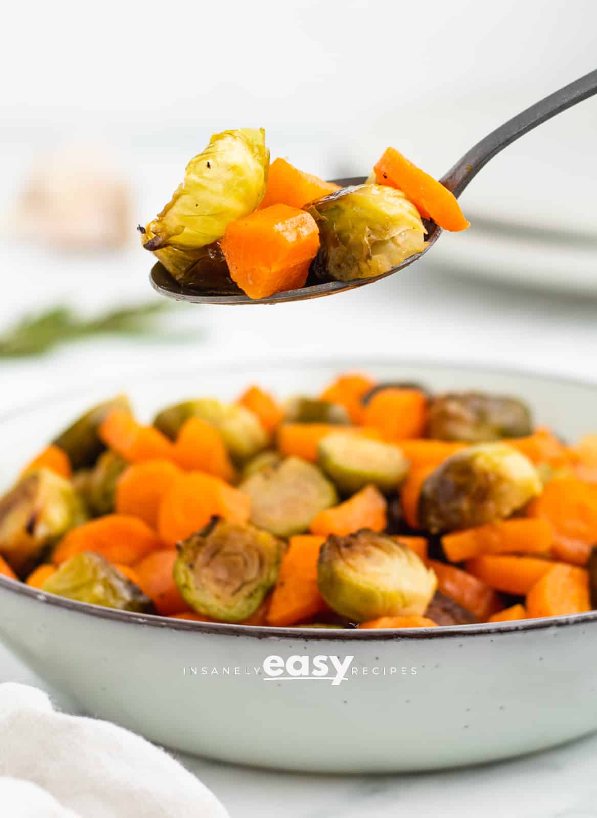 roasted brussel sprouts in a white bowl with a silver spoon above the bowl full of carrots and brussels