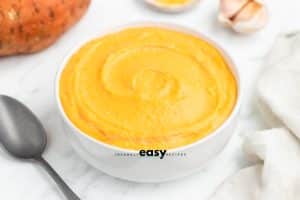 photo of sweet potato sauce in a white bowl, with a spoon and a white kitchen towel in the foreground, and a sweet potato and garlic head in the background