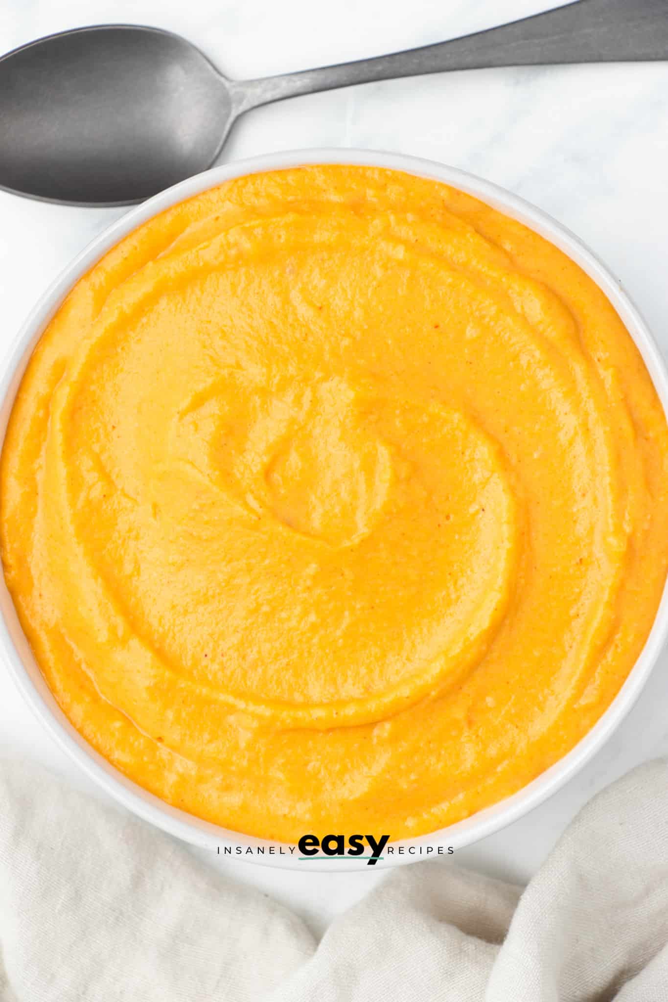 top view photo of sweet potato sauce in a white bowl with a spoon above it and a white kitchen towel below it