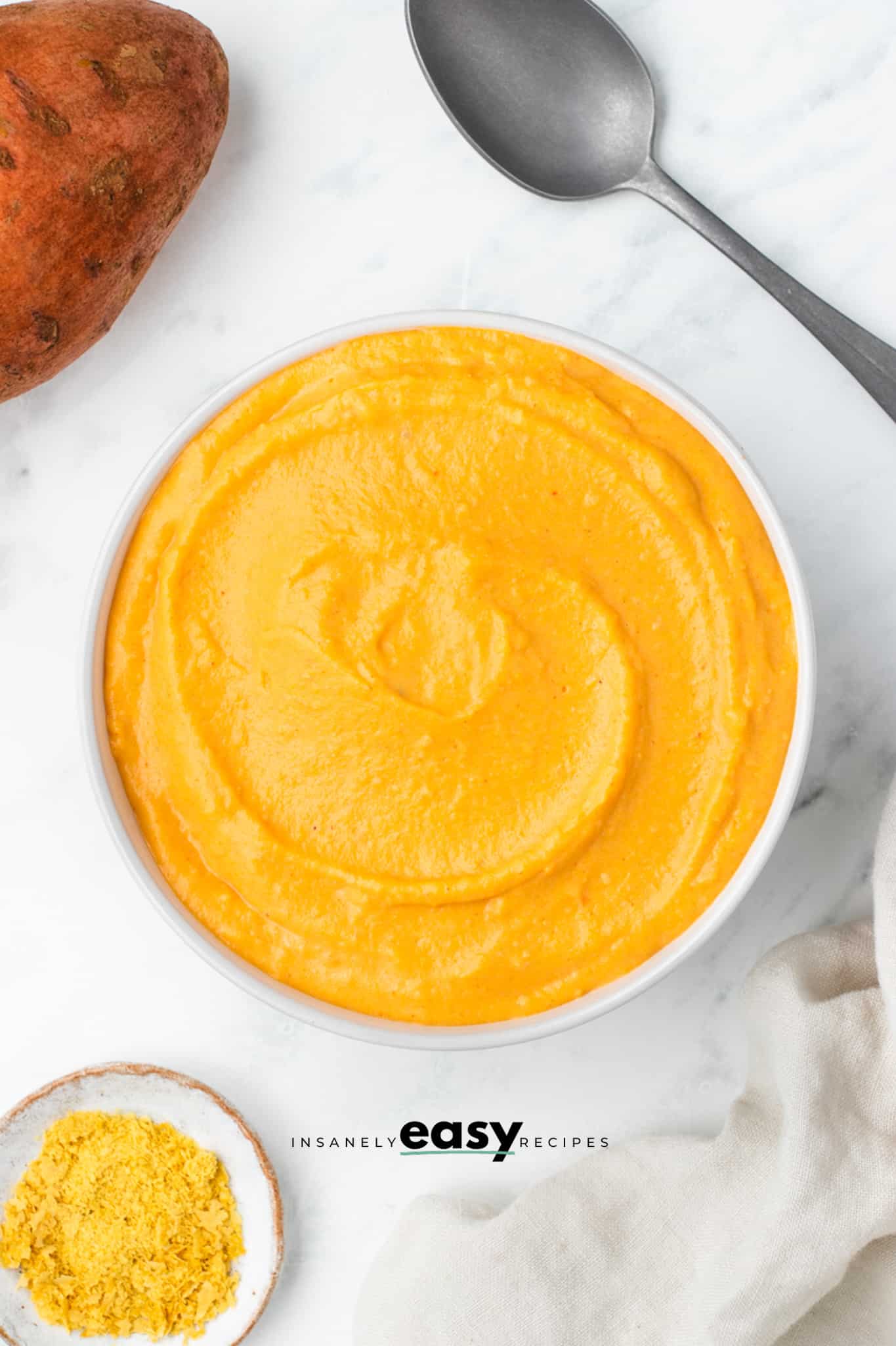 top view photo of sweet potato sauce in a white bowl with a spoon and a sweet potato above it and a white kitchen towel and small white bowl of turmeric below it