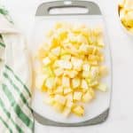diced apples on a white and gray cutting board a bowl of apples to top right and green and white town to the left