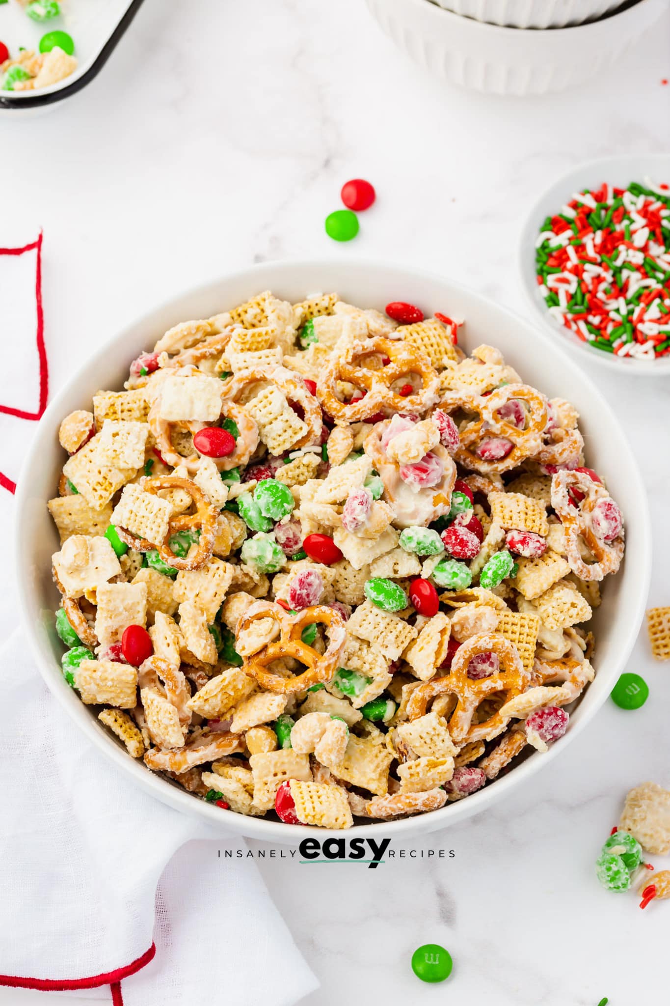 a round white bowl filled with holiday white chocolate chex mix. Sprinkles and M&ms are strewn around.